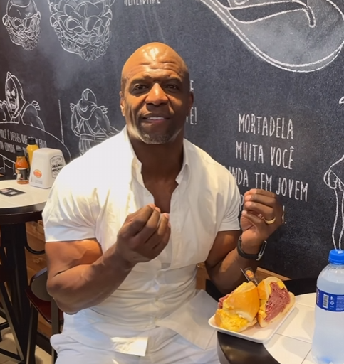 Ator Terry Crews sentado ao lado do famoso pão de mortadela do Mercadão de SP